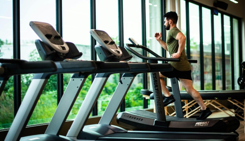 Young man workouts on treadmill in modern gym with large windows and natural light.