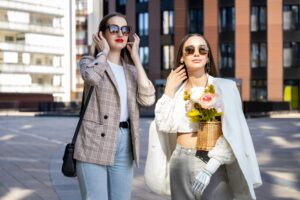 Two stylish women enjoy a sunny day in the city wearing chic outfits and sunglasses.